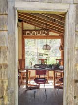 Dining room with large table, large window, and slanting ceiling with exposed beams is seen through open doorway of shingled house in Concrete, Washington, designed by Jes Paone.