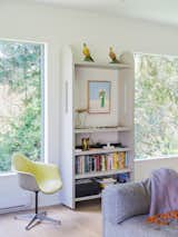 Living room of home in Concrete, Washington, designed by Jes Paone with white walls and ceiling, light hard wood floors, grey sofa, pale yellow armchair, white wood storage cabinetry bookshelf, and framed views of surrounding forest.