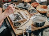 Woman uses tongs to place different minerals on wood block covered in cement paste at a desk workbench in an art studio with arched curving roof built on a flatbed trailer in Salida, CO.