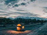 An art studio with arched curving roof built on a flatbed trailer in Salida, CO, sits parked a dry desert arid mountainous terrain landscape beside train tracks at dusk.