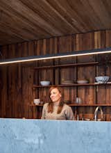 Woman looks out from behind marble kitchen island in a prefabricated home in Bariloche, Argentina, by Nicolás Tovo and Teresa Sarmiento, founders of the architecture and design studio La Base with dark toned wood clad walls, long metal pendant lights, open shelving stacked with ceramicware, and light-toned wood ceiling.