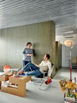 Man sits in metal armchair upholstered in black leather as he talks to partner leaning on floor-to-ceiling green-tinged MDF partition in an apartment in Madrid, Spain, renovated by architect Eduardo Mediero with metal slatted ceiling, white rubber flooring, blockish geometric coffee table, assorted floor and table lamps, and rectangular pendant lights.