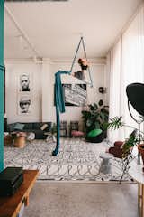 Woman practices aerial dancing with blue cords ribbons suspended from hook in ceiling of living room in a factory converted into an apartment by Gabriele Schiavon, creative director of Barcelona-based design studio Lagranja with white and black geometric area rug, floor-to-ceiling windows, blue-grey sofa, large potted plants, and white walls and ceilings.