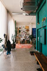 A factory converted into an apartment by Gabriele Schiavon, creative director of Barcelona-based design studio Lagranja with brick walls painted dark blue green turquoise, long medium-toned wood bench, terrazzo floors, tall potted cactus, exposed vents, hexagonal orange-and-white area rug, and double-height ceilings.