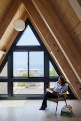 Woman reads on armchair upholstered in pale blue fabric set beside a stack of books in lofted space beneath slating roof in a 1960s A-frame with in Manhattan Beach, Los Angles, CA, renovated by Julie Anderson and Amy Dantzler in collaboration with architect Bryan Libit.