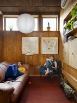 Man lays on dark pink couch as woman reads a magazine in a dark wood armchair upholstered in turquoise fabric in living room with red pink area rug, large globe parchment pendant light, cork and wood-paneled walls, cork-paneled ceiling, and framed cartographic artwork in a house by architect Jack Becker and landscape architect Maddie Hoagland-Hanson in an alley in the Capitol Hill area of Washington, D.C.