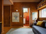 Livingroom in a house by architect Jack Becker and landscape architect Maddie Hoagland-Hanson in an alley in the Capitol Hill area of Washington, D.C. with grey-green area rug, blue-grey sofa with yellow cushion, sculpture of a goose, medium-toned wood paneled walls, cartographic artwork, cork flooring, exposed wood shelving nestled between ceiling and long window, and open doorway leading into bath-room with red-tiled floors, walls lined with medium-toned wood and red-tiling, and a pedestal sink sitting on a white vanity.