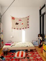Girl walks into her bedroom in a converted loft apartment in Green Point, New York City, renovated by interior stylist and designer Merie Subryan with thick pink-red rug woven with multicolored diamond pattern, low plywood bed with wheels, tapestry quilt hanging on a white wall, toys and books stowed in bookcase and in bins beneath the bed, yellow wireframe storage unit, and black framed acrylic screen separating it from the adjacent nursery and office.