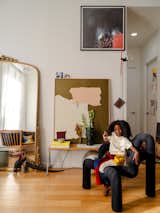 Girl sits on black chair in the living room of converted loft in Green Point, New York City, renovated by interior stylist and designer Merie Subryan with light wood floors, large mirror set in ornate gold brass frame, low metal bench with light-toned wood seat, and contemporary abstract artwork.