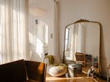 Living room of converted loft in Green Point, New York City, renovated by interior stylist and designer Merie Subryan with light wood floors, plush white sofa chair, grey end table, light-toned wood sofa chair with brown upholstery, and large mirror with ornate gold brass frame. 