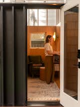 Woman works at standing desk in office with plywood interior, white board, velvet green armchair, brown stool ottoman, high polycarbonate clerestory windows, built-in storage, and round flat green wall light sconce in work shed by Linework Architecture in backyard of home in Seattle, Washington.