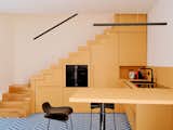 Bright yellow-orange kitchen beneath staircase with grey-blue herringbone tile floors and kitchen island surrounded by low-backed black bar stools in a tiny seaside home for Massimo Ricci and Valentina Martinelli in Termoli, Italy, renovated by Rispoli Guerrera Architetti.