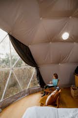 Child eats snack while sitting on a medium-toned wood chair beside triangular dark coffee table sits beneath cushiony insulation in a geodesic dome by FDomes with an interior designed and built by SpotLab in Lost Woods glamping site situated in Boyne Falls, Northern Michigan.