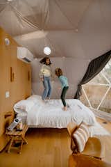 Children jump on bed sitting against white oak millwork unit with rounded edges beneath cushiony insulation in a bedroom with night stand, light hardwood floors, medium-toned wood chairs with brown-and-white cushions, and projector in geodesic dome by FDomes with an interior designed and built by SpotLab in Lost Woods glamping site situated in Boyne Falls, Northern Michigan.