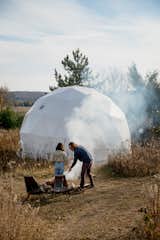 Couple tend bonfire outside white geodesic dome by FDomes with an interior designed and built by SpotLab in forested Lost Woods glamping site situated in Boyne Falls, Northern Michigan.