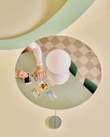 Seen through a reflection of a flat metal pendant light hung from a pastel yellow and green ceiling, a woman sorts through tarot cards on a past green Formica topped dining table in a room with brown checkered flooring in in an apartment in New York City’s The Belnord building, featured as The Arconia in Only Murders in the Building, renovated by New York designers Talitha Liu and Lexi Tsien.