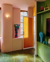TV set into green nook with rounded edges of pink millwork unit beside open metal-framed pink door set with bumpy yellow bottle glass in an apartment in New York City’s The Belnord building, featured as The Arconia in Only Murders in the Building, renovated by New York designers Talitha Liu and Lexi Tsien.