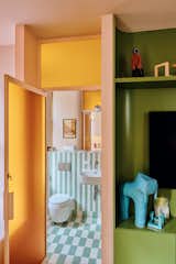 Past a pink and olive-green millwork unit with rounded edges and through a pink metal-framed door set with yellow bumpy bottle glass is a bathroom lined with mint and white tile arranged in checkered and striped patterns, a one-piece wall mounted toilet, and wall mounted sink in an apartment in New York City’s The Belnord building, featured as The Arconia in Only Murders in the Building, renovated by New York designers Talitha Liu and Lexi Tsien.