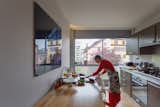 Women prepares meal at long wood table in long and narrow kitchen with dark brown cabinetry, metal counters, and floor-to-ceiling glass window set into short wall in an apartment in the prototypical urban residential apartment building the Immeuble Clarté in 1932, a now UNESCO World Heritage site in Geneva, Switzerland commissioned by Edmond Wanner and designed by Le Corbusier and his cousin Pierre Jeanneret.