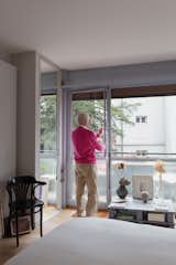 Man opens glass door in bedroom of an apartment in the prototypical urban residential apartment building the Immeuble Clarté in 1932, a now UNESCO World Heritage site in Geneva, Switzerland commissioned by Edmond Wanner and designed by Le Corbusier and his cousin Pierre Jeanneret with black armchair, low wooden grey coffee table, and floor-to-ceiling glass windows.