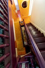 Purple-toned staircase in a Victorian home in Providence, Rhode Island, renovated by Emily Lindberg Design and Work in Use rises from second-story hallway with long mustard rug with abstract floral embroidered motifs, bathroom with red-and-white wavy tiles, and hardwood floors, with some wall clad with wood slats and others painted white.
