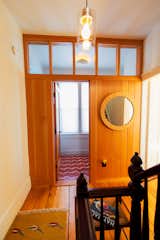 In a Victorian home in Providence, Rhode Island, renovated by Emily Lindberg Design and Work in Use, the door to a bathroom on the second floor stands ajar, providing a view of the large translucent windows and red-and-white wavy tile within. A round mirror hangs on the wood-clad wall portioning the bathroom from the hall. Translucent clerestory windows are set above the door.