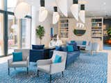 Large room with blue and white carpet with abstract ocean pattern, large pendant lamps, long blue velvet sofas, grey velvet armchairs, large windows with grey trimming looking out into central courtyard, and light-toned wood millwork unit with large round mirror and shelves laden with books and ceramics in a midcentury, neo-Breton post office building designed by architect Peirre-Jack Laloy and renovated into senior housing by Paris firm Chatillon Architectes in Brest, France.