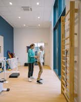 Older woman receives physical therapy in room with light-toned wood stretching ladder mounted onto wall, light-toned wood floors, floor-to-ceiling windows with blue trimming, and therapeutic equipment in a midcentury, neo-Breton post office building designed by architect Peirre-Jack Laloy and renovated into senior housing by Paris firm Chatillon Architectes.