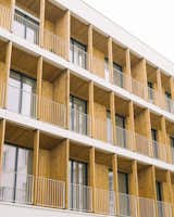 Rows of recessed balconies with walls and ceilings clad in yellowish medium-toned wood slats and tall sliding glass doors with dark grey trimming look out over courtyard in a midcentury, neo-Breton post office building designed by architect Peirre-Jack Laloy and renovated into senior housing by Paris firm Chatillon Architectes.