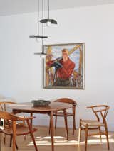 Round medium-toned wood dining table and armchairs sits beneath globe pendant lights with flattened black leaf-shaped fan-shaped shades beside painting of skier in vintage gear in dining room with light-toned hardwood floors and white walls and ceiling in a prefab home designed by architect Gabriele Mary Ann Schicketanz built from modular units by Method Homes in Carmel, California.