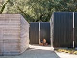 A boy rests with a dog on a wooden bench on a gravel path between the walls of a prefab home designed by architect Gabriele Mary Ann Schicketanz built from modular units by Method Homes in Carmel, California, with yakisugi blackened wood cladding.
