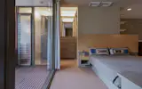 The airy primary bedroom is open on both sides. A large glass slider provides access to a balcony, and a low wall of shelving allows views over the living room.