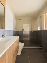 Dark-gray, 24-by-24-inch ceramic tile wraps the primary bathroom as wainscoting. In the shower, the tiled lower wall projects outward to form a ledge for soap and shampoo. The mirror surround and vanity cabinet are in the same white oak used throughout the house.