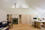 Another view of the living area shows a green-tiled fireplace and the kitchen’s island and cabinets, made of whitewashed birch plywood. The vintage stools are by <a href="https://www.2modern.com/products/high-stool-64-counter-height?variant=8267400380461&amp;utm_source=google&amp;campaign_id=13898193351&amp;ad_id=533579341404&amp;utm_medium=cpc&amp;utm_campaign=X99.%20All%20products%20-%20Shopping%20[SHOPPING]&amp;utm_content=123152861005&amp;utm_term=&amp;gclid=Cj0KCQjwt9jVBhDXARIsAFSP-6dO-anEq7NzyGs5J2lbUMhhpi5RjaRkF7lRSM5zObeZhdYrntOUGXUaAt2tEALw_wcB&amp;gad_source=1&amp;gad_campaignid=13898193351&amp;gbraid=0AAAAAD_WqgnDnIXb8-y05VCitcHRhuqdD">Artek</a>.