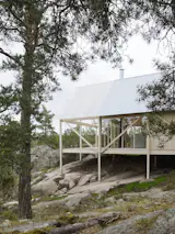 Viggsö is a minimal residence located in Värmdö, Sweden, designed by Arrhov Frick Arkitektkontor. A landscape of stone cliffs, wind bent pine trees, heather, and a forest floor of lichen and wild berries – this sparse yet rich site, coupled with a minimal budget, establishes the principles for this private residence at its origin. The design process largely focused on the development of a simple and economic structure in wood. The materiality of the house had to be efficiently transported and assembled on the island, yet able to withstand relatively large spans. The foundation, a series of posts, minimizes the houses touch on the ground condition, while elevating it to sit amongst the trees and vegetation. Simple planks connect the residence with the surrounding cliffs, following the aim to create an unassuming space within the landscape.