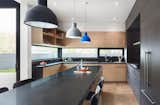 The contrasting material palette selected for the house is seen in the selection of finishes used in the kitchen, including white oak and charcoal cabinetry and dark stone counters. A long horizontal window provides framed views of the backyard, further emphasizing the connection between the interior and exterior spaces.