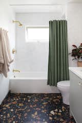 In the kids' bathroom, Marmoreal flooring with a black background and green, gold, and green spots, a Nickey Kehoe-designed green-and-white shower curtain, pale green Farrow &amp; Ball vanity paint, and brass fixtures add color and playful touches.