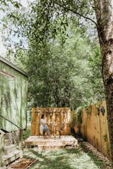 A small deck is shaded by towering trees in the fenced front yard of one of the tiny homes.