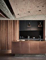 In the open kitchen, wood-paneled sliding doors conceal dark cabinetry, and together with a sleek, dark counter, and a shiny bronze-clad island with a sink, the kitchen becomes part of the overall design rather than simply an area of utility.
