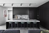 B.E Architecture combines a revitalized kit home with a modern steel-and-glass extension to form a multi-generational Melbourne residence. In the kitchen, black cabinets meld seamlessly with dark countertops, furniture, and sleek track lighting.