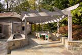 A new flagstone patio protects guests from the sun while relaxing or dining. A large pergola is bordered by a gas fire pit for warmth and ambient lighting. Pendant lights hang from the pergola for additional light after sunset.