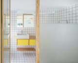 The bathroom has two glass walls with frosted and transparent glass to allow light to filter in from the windowed area. Colored grout and yellow drawers and hardware bring bright colors into the space.