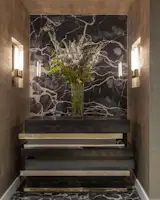 The entryway features a bronze, stainless steel, and wood console against a backdrop of precious stone. The walls and ceilings were given a faux finish of metallic woven, grass-cloth type texture, bringing an element of glamour and drama.