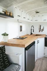 The countertops are birch-wrapped plywood. The matte black hardware and faucet punctuate white cabinets and peel-and-stick tile. A magnetic knife strip and mounted paper towel holder is another way they can save space. The dish rack is folded and stored under the sink when not in use.
