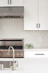 A kitchen is kept light and bright with white cabinets with dark hardware, white countertops, and a geometric patterned backsplash in a range of whites, creams, and beiges from Dwell patterns Heath tile backsplash.