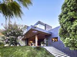 From the backyard's lower level, rectangular forms extend towards the view with a glass cube at the upper floor. The roof overhang with a white oak lid extends from the living room to the exterior. An oculus gives a peek to the garden roof from below. A brass scupper stretches to throw excess irrigation water of the above roof garden to a circular, steel catch basin that rises above the no-mow fescue grass of the hillside. Concrete steps lead to the garage of the lower level. An understanding of this street's ridgetop shape is partially understood from this view:  The ridge slowly falls to the North (San Fernando Valley behind the viewer).