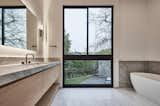 Primary bath features a view of the expansive yard. Two single vanities - one at the west end and the other at the east end (this view) of the bathroom are bi-parted by the entrance to the walk-in closet. Sconces by Kelly Wearstler and plumbing fixtures by Dornbracht.