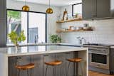 Counter stools tuck into the extra deep kitchen island. Floating shelves made from slabs of white oak run between the tile backsplash. The kitchen is fitted with a full Fisher & Paykel suite of appliances as well as French gray shaker panel cabinetry and restoration hardware cabinetry pulls and knobs. Clerestory windows allow for south-facing natural light exposure throughout the day