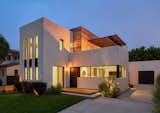 Front yard at dusk with glow from second floor deck outdoor firepit - layered planes of smooth white plaster and wood siding punctuated by dark bronze windows and doors. Inspired by minimalism and Art Deco.