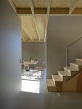 Interior view from guest room to staircase and kitchen. The staircase is made of pine with a sliding rack attached to the catch. The roof with visible solid wood construction and walls of grey coloured wood fibreboards. The floor is glossy and dust-bound concrete floor.